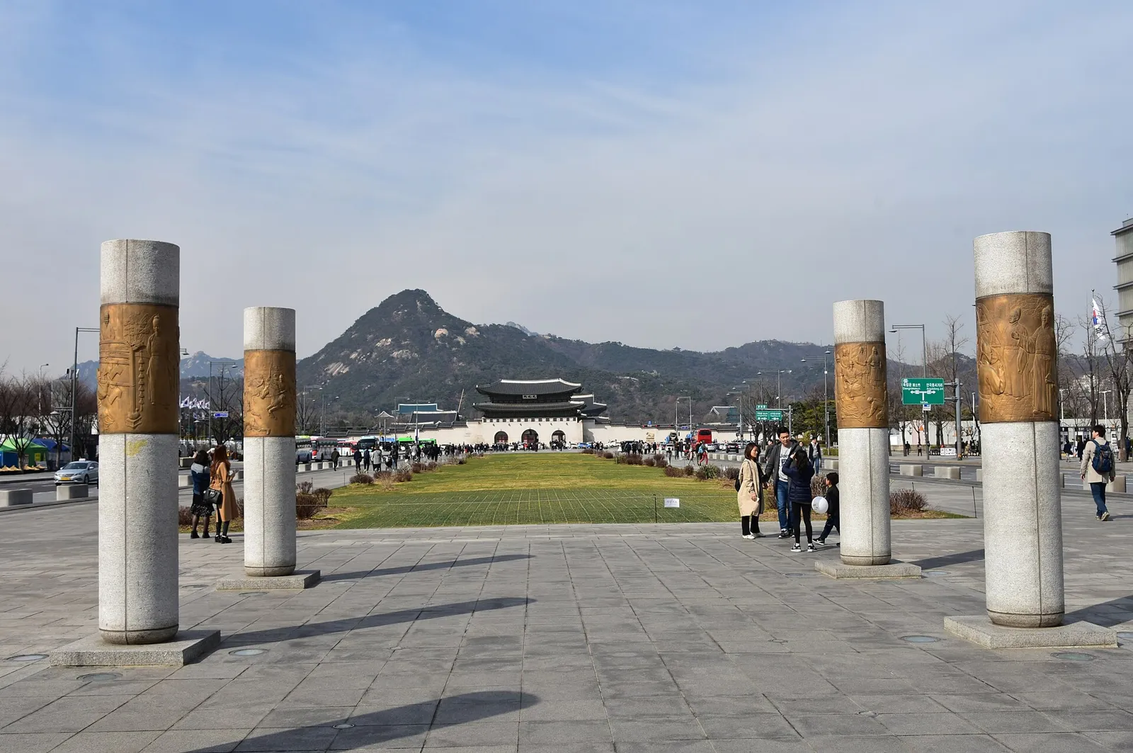 맑은 날 서울 광화문광장 전경 실사진: 앞쪽에 부조가 새겨진 석주 4개, 가운데 잔디 광장, 멀리 광화문과 북악산, 산책하는 사람들