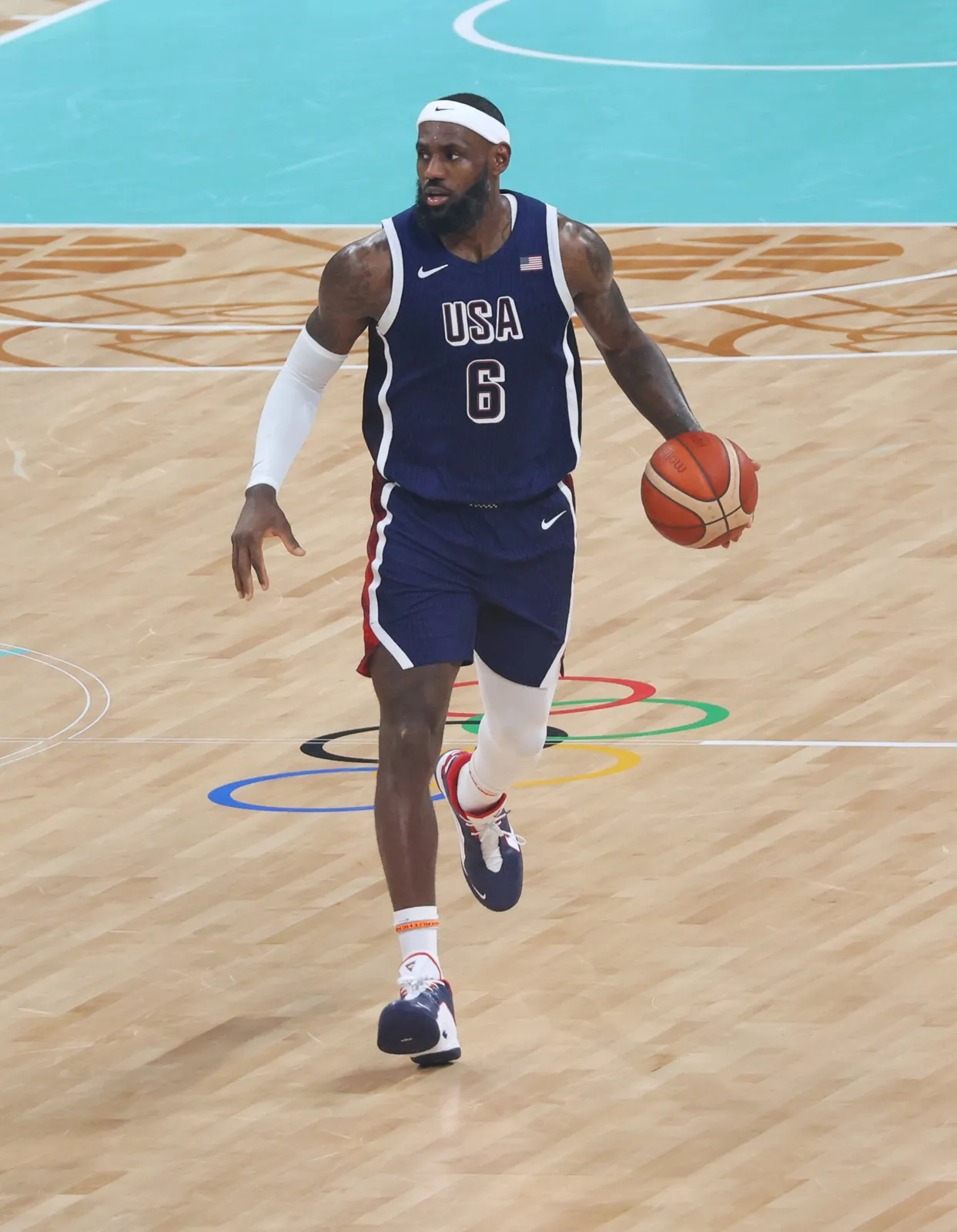 네이비색 'USA' 6번 유니폼에 흰색 헤드밴드를 한 농구 선수가 올림픽 오륜 로고가 그려진 코트에서 드리블하는 실사진