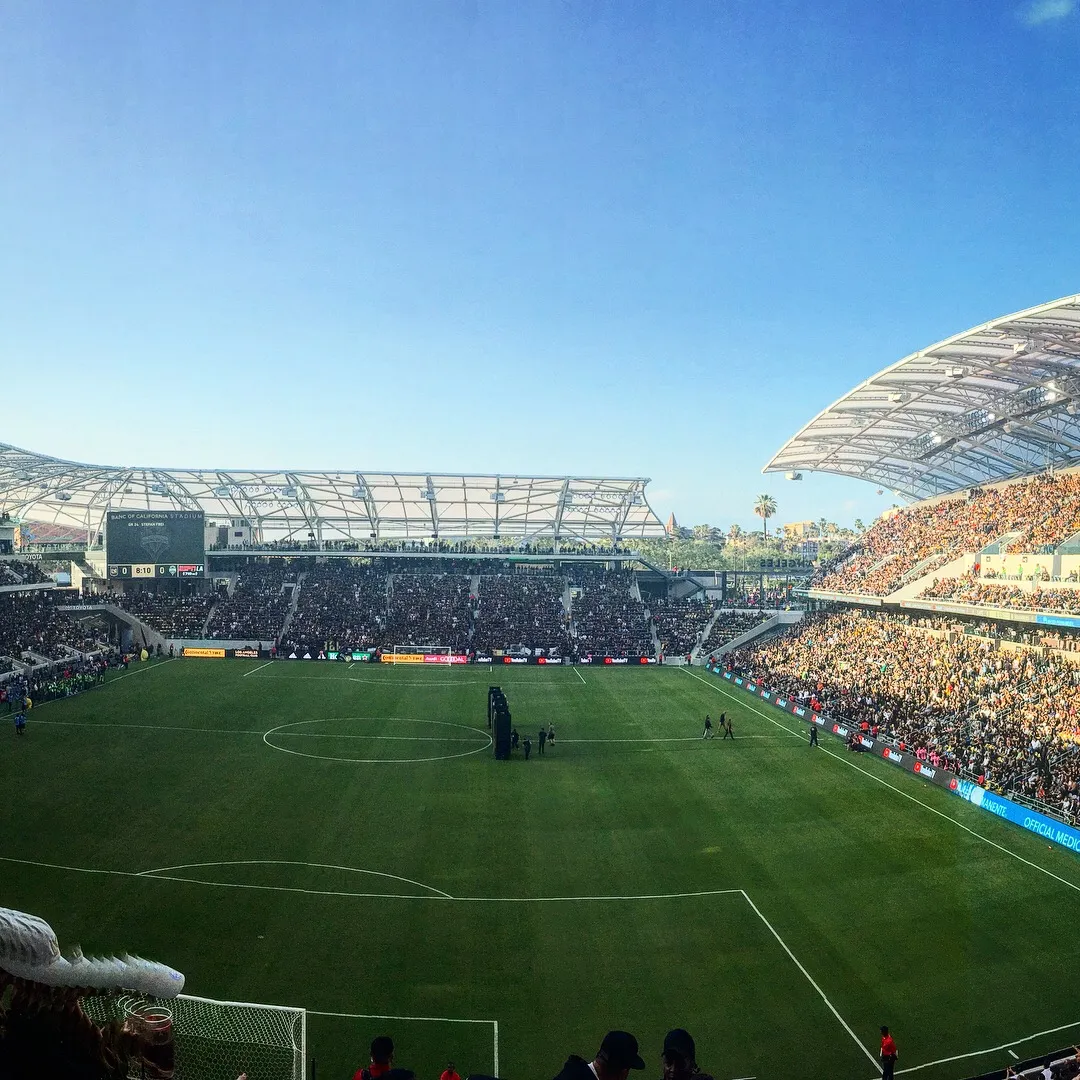 해가 밝은 낮, 관중이 들어찬 축구 경기장의 전경 파노라마. 전광판에 'Banc of California Stadium'이라는 글자가 보이고, 잔디 위에는 소수의 사람이 서 있다. 특정 인물을 알아볼 수 있는 장면은 아니다.