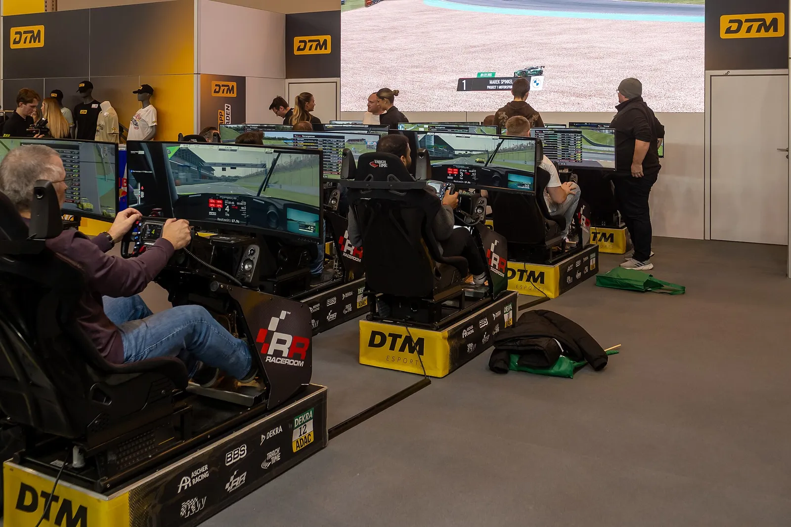실내 전시장에서 여러 사람이 DTM Esports 로고가 붙은 레이싱 시뮬레이터 좌석에 앉아 모니터로 레이싱 게임을 하고 있고, 뒤쪽 대형 화면에도 레이싱 게임 화면이 나온 실사진