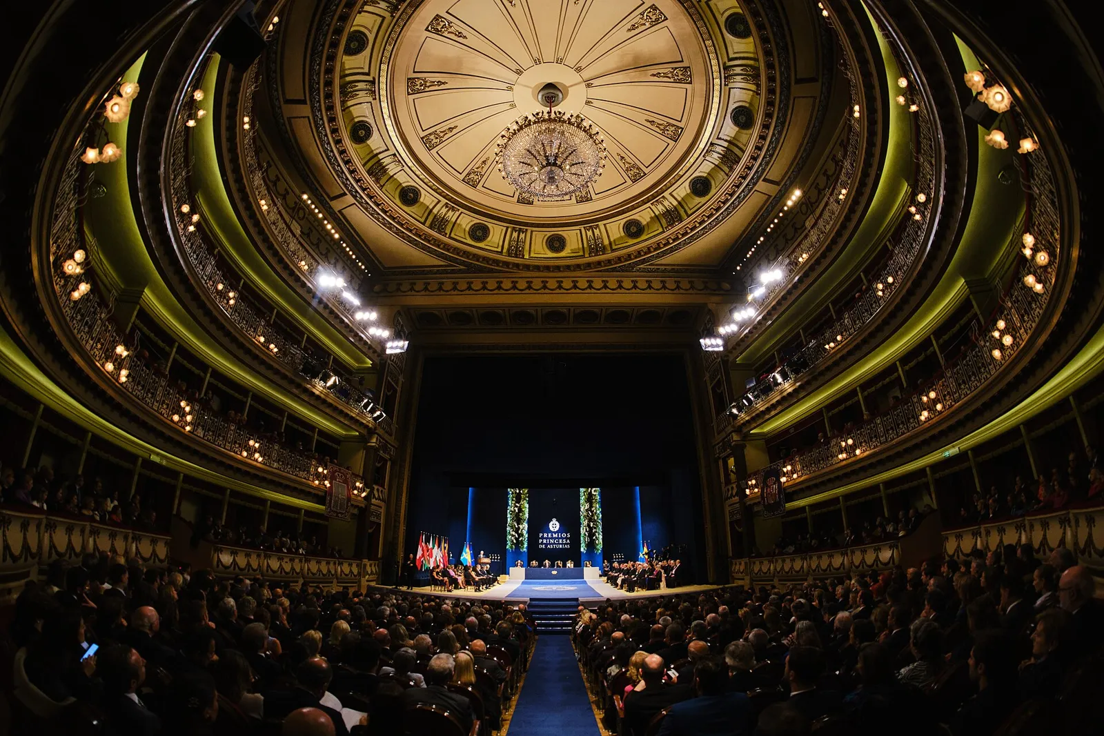 청중이 가득 찬 고풍스러운 극장 내부 광각 사진. 무대 뒤 배경막에 'PREMIOS PRINCESA DE ASTURIAS' 로고와 깃발, 꽃장식이 보이고 단상에 인사들이 앉아 있다.