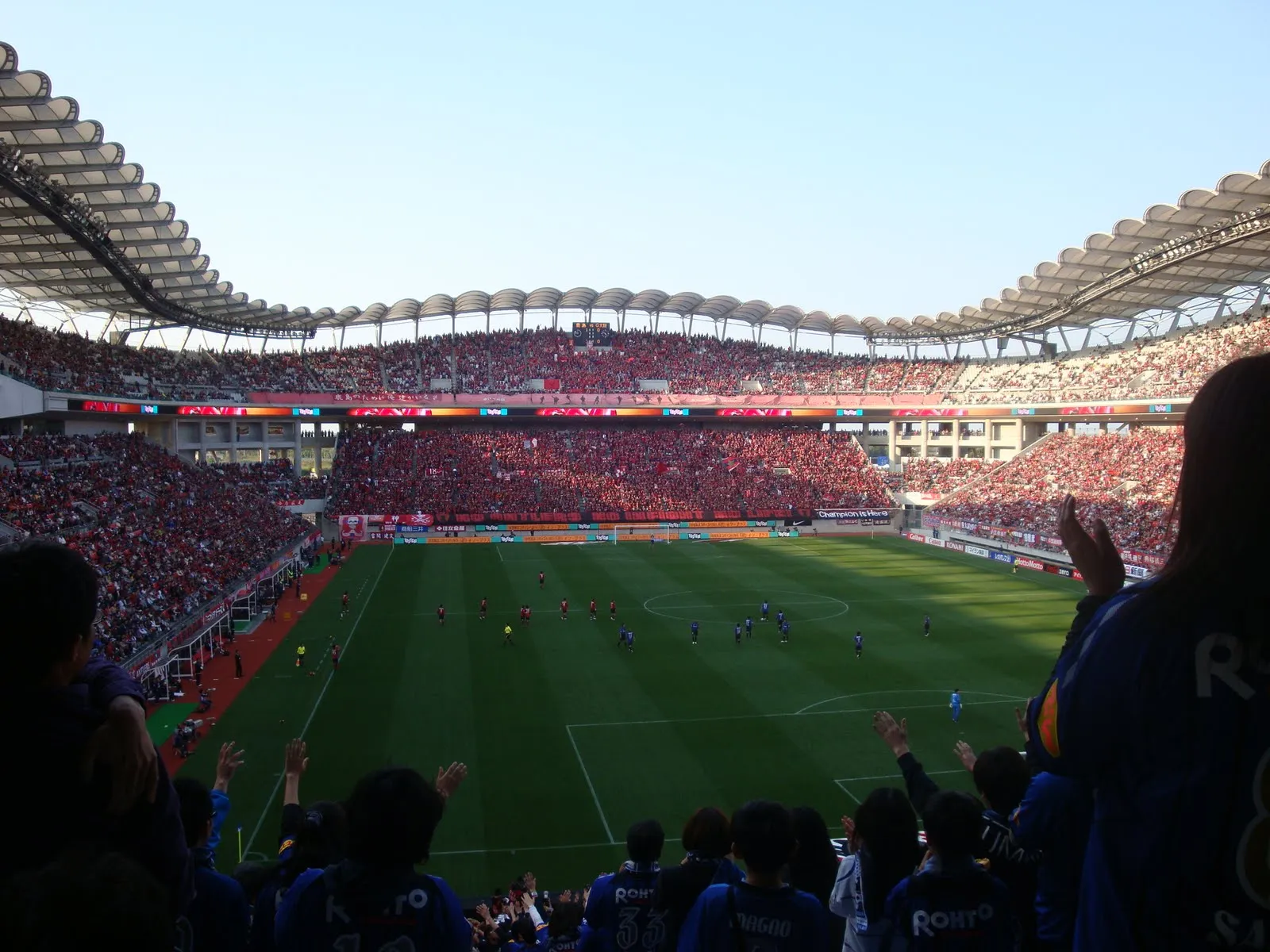 관중석이 붉은 옷의 팬들로 가득 찬 물결형 지붕 구조의 대형 축구장 전경이다. 잔디 위에 선수들이 있고, 전경에는 파란 유니폼을 입은 원정 팬들이 손뼉을 치고 있으며, 멀리 스탠드 중앙에 전광판이 작게 보인다.